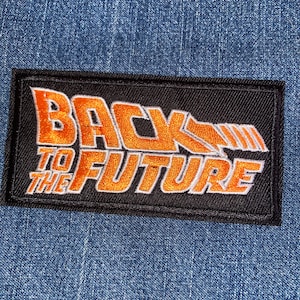 May include: A rectangular black patch with the words "BACK TO THE FUTURE" embroidered in orange and white. The patch is set against a blue denim background, showcasing the iconic movie title.