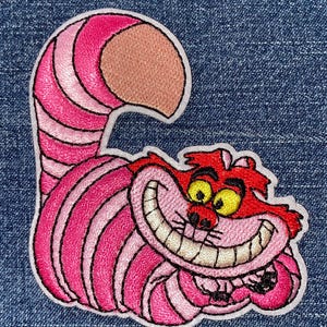 May include: An embroidered patch featuring a stylized cat with a wide, toothy grin. The cat has bright pink and dark pink striped fur, a large curved tail, and yellow eyes. The design is outlined in white thread against a blue denim background.