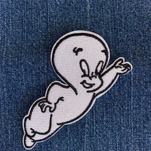 May include: A white cartoon ghost with a black outline, smiling and waving. The ghost is flying in the air. The image is on a blue denim background.