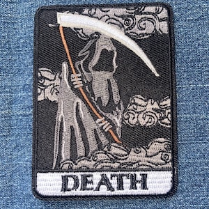 May include: A rectangular embroidered patch featuring a black background with a grey figure of a hooded skeleton holding a scythe. The design includes grey swirling clouds and a white bottom border with the word DEATH embroidered in black letters.