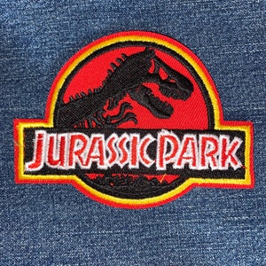 May include: Embroidered patch featuring the Jurassic Park logo. The patch has a red background with a black dinosaur silhouette and yellow and white accents. The text "JURASSIC PARK" is displayed in white letters with a red outline.