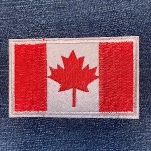May include: Embroidered patch featuring the Canadian flag. The design includes a red maple leaf on a white background, flanked by red vertical stripes. The patch has a white border and is rectangular in shape.