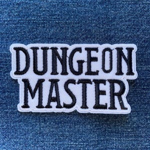 May include: A white patch with black text that reads "DUNGEON MASTER". The patch has a black border and is set against a blue denim background. The patch is likely for clothing or accessories.
