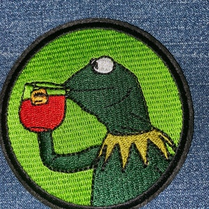May include: Green felt embroidered patch of a frog drinking from a red cup. The frog has a yellow crown and a white eye.