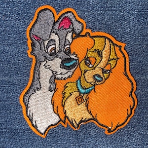May include: Embroidered patch featuring the characters Tramp and Lady from the animated film "Lady and the Tramp." The patch has an orange border, with Tramp in gray and Lady in shades of orange and gold. Lady wears a blue collar.