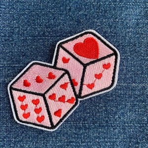 May include: Two pink dice with red hearts embroidered on each side. The dice are sewn together and have a white outline.