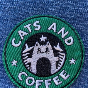 May include: Embroidered patch with a green border and the words "CATS AND COFFEE" in white. The design features a cat in the center, surrounded by a black circle and white stars. The patch is on a blue denim background.