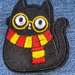 May include: An embroidered patch featuring a black cat with large white eyes and round yellow glasses. The cat is wearing a scarf with horizontal red and yellow stripes. The patch has a black stitched border and a textured fabric surface.