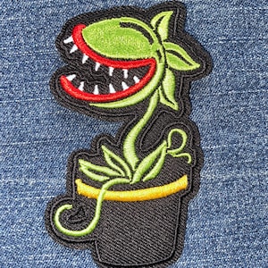May include: Embroidered patch featuring a cartoon plant with a large, open red mouth filled with white teeth. The plant has green leaves and a stem, and is in a black pot with a yellow rim. The patch is on a blue denim background.