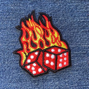 May include: Embroidered patch featuring two red dice with white dots, set against a backdrop of yellow and red flames. The patch has a black border and is placed on a blue denim fabric.