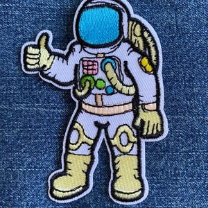 May include: Embroidered patch of an astronaut in a white spacesuit with a blue visor, giving a thumbs-up. The patch features yellow gloves, boots, and accents, with a black outline. The astronaut's backpack and suit details are visible.