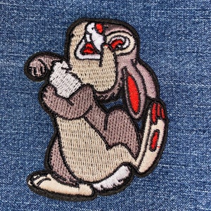 May include: A brown and gray embroidered patch of a cartoon rabbit with red accents. The rabbit is sitting on its hind legs with its front paws raised to its face. The rabbit has a surprised expression.