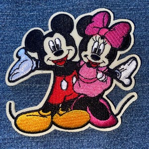 May include: Embroidered patch featuring Mickey Mouse and Minnie Mouse. Mickey is wearing red shorts and yellow shoes, while Minnie is in a pink dress and bow. The patch has a white border and is set against a blue denim background.