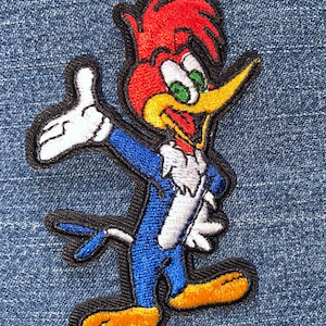 May include: Embroidered patch featuring Woody Woodpecker in a classic pose. The patch showcases the cartoon character with a red head, yellow beak, blue body, and white gloves. The patch is outlined in black.