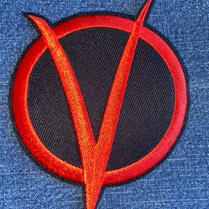 May include: Embroidered patch featuring a red 'V' inside a red circle, set against a black background. The patch is sewn onto a blue denim fabric, creating a striking contrast. Ideal for jackets, bags, or other apparel.