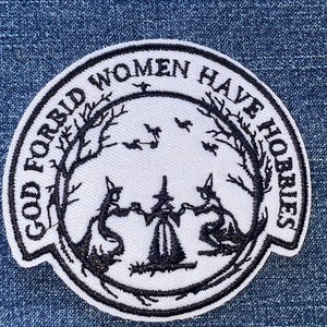 May include: A circular embroidered patch with a white background and black stitching. The design features three figures in pointed hats holding hands, surrounded by bare trees and flying bats. The text reads GOD FORBID WOMEN HAVE HOBBIES.