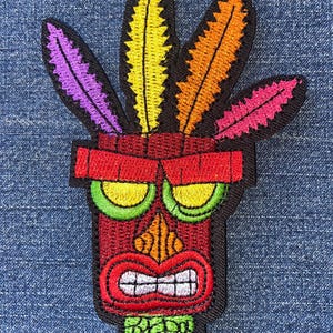 May include: Embroidered patch featuring a stylized tiki mask with a red face, green eyes, and a wide, toothy grin. The mask has colorful feathers in yellow, orange, pink, and purple. The patch is outlined in black.
