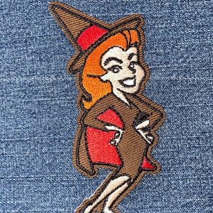 May include: Embroidered iron-on patch featuring a cartoon witch with brown hair, a brown dress, a red cape, and a brown witch hat.
