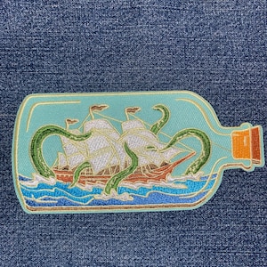 May include: Embroidered patch featuring a ship inside a bottle, with green tentacles reaching out. The ship has white sails and brown accents, set against a light blue background with blue waves. The bottle's rim is brown.