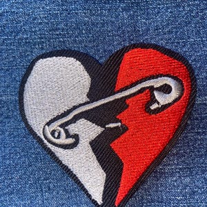 May include: Embroidered patch featuring a broken heart design. The heart is split into two halves, one white and one red, with a silver safety pin going through the break. The patch is on a blue denim background.