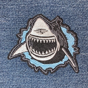 May include: Embroidered patch featuring a cartoon shark with an open mouth, showcasing sharp teeth. The shark is black, white, and gray, set against a light blue background, with a torn-edge design. The patch is on a blue denim background.