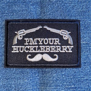 May include: A black embroidered patch with a white outline. The patch features two crossed pistols, a mustache, and the text "PM YOUR HUCKLEBERRY".