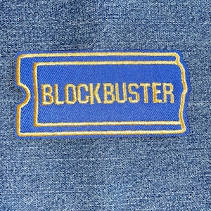 May include: A blue embroidered patch in the shape of a movie ticket, with the word "BLOCKBUSTER" in gold lettering. The patch has a gold border and is set against a denim background.