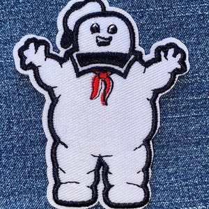 May include: An embroidered patch featuring a white, marshmallow-like character with a black outline. The figure wears a black sailor collar and a red neckerchief, with a small black hat perched on its head, arms raised in a welcoming gesture.