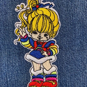 May include: Embroidered patch featuring a cartoon character with yellow hair, wearing a blue dress, and colorful striped sleeves and leggings. The character's red shoes have yellow soles. The patch is outlined in white and set against a blue denim background.