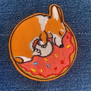 May include: Embroidered patch featuring a corgi dog curled up on a donut. The corgi is orange and white, the donut has pink frosting with colorful sprinkles, and the patch has a gold border. The patch is on a blue denim background.