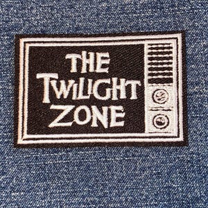 May include: A black and white embroidered patch featuring the text "THE TWILIGHT ZONE" in a stylized font. The patch has a rectangular shape with a white border and a retro television design on the right side. The patch is set against a blue denim background.