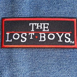 May include: A rectangular black patch with the words "THE LOST BOYS" in white lettering. The patch has a red border and is set against a blue denim background. The text includes a small red dot.