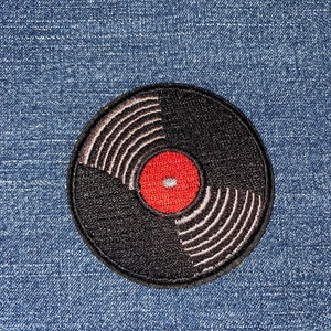 May include: Embroidered patch featuring a black vinyl record design with white grooves and a red center. The patch is circular and has a detailed, textured appearance, suitable for application on clothing or accessories.