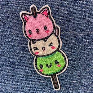 May include: A pink, white, and green embroidered patch of three cute cartoon cats on a stick. The cats have smiling faces and are stacked on top of each other.