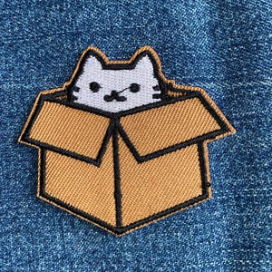 May include: Embroidered patch featuring a gray and white cat peeking out of a brown cardboard box. The box has black outlines and is stitched onto a blue denim background. A cute, whimsical design.