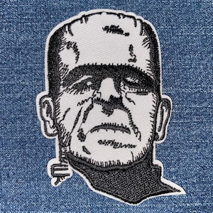 May include: Embroidered patch featuring a black and white illustration of Frankenstein's monster. The patch depicts the monster's face with detailed stitching, including facial features and a black outline. The patch is set against a blue denim background.
