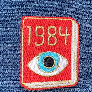 May include: A rectangular embroidered patch featuring a bright red book cover with the text 1984 stitched in gold thread. Below the text is a detailed eye graphic with a black pupil, a vibrant blue iris, and white sclera, all outlined in gold.