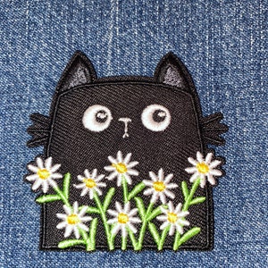 May include: Black cat patch with white daisy flowers and green stems. The cat has large white eyes and small ears. The patch is on a blue denim background.