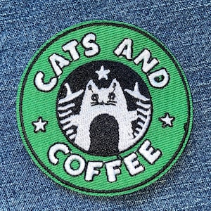May include: A circular embroidered patch with a bright green border and a black center. The text "CATS AND COFFEE" is stitched in white around the perimeter. A white stylized cat illustration sits in the middle with two small stars.