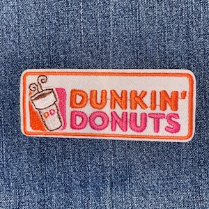 May include: A rectangular patch featuring the Dunkin' Donuts logo. The patch has a white background with an orange border. The words "DUNKIN' DONUTS" are in orange and pink lettering. A coffee cup graphic is on the left.
