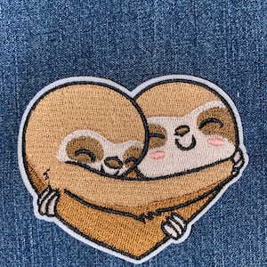 May include: Embroidered patch featuring two sloths hugging in a heart shape. The sloths are light brown with darker brown accents and smiling faces. The patch has a white border and is set against a blue denim background.
