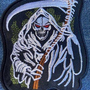 May include: Embroidered patch featuring a white skeletal figure in a hooded robe, holding a scythe with a brown handle. The figure has red eyes and is set against a black background with green and white accents.