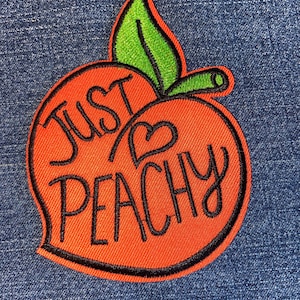 May include: Orange peach-shaped patch with black stitching and the text "Just Peachy" in black cursive lettering. The peach has a green leaf on top.