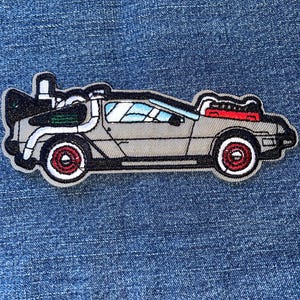 May include: Embroidered patch featuring a gray DeLorean car with red wheels and accents. The patch has a black outline and a white border. The car's details include windows, vents, and a rear engine. The patch is likely for clothing or accessories.
