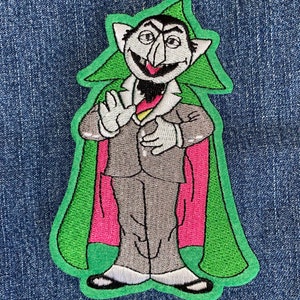 May include: Embroidered iron-on patch featuring a green-skinned, red-caped, and gray-suited monster with a large nose and a wide smile. The monster is wearing a green hat and has a black beard.