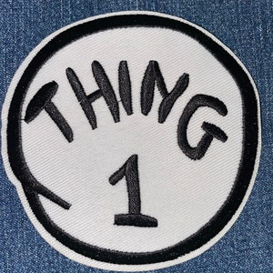 May include: White round patch with a black border. The patch has the word "THING" written in black cursive lettering and the number "1" in black below it.
