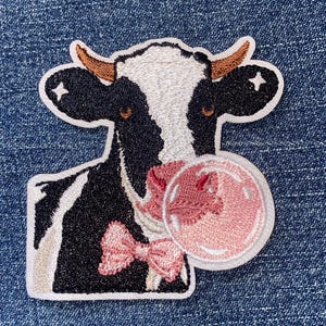 May include: An embroidered patch featuring a black and white cow with brown horns, wearing a pink bow tie and blowing a large pink bubblegum bubble. The cow has small white star accents on its ears and is outlined in white thread.