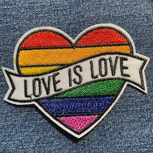 May include: Embroidered patch in the shape of a heart with a rainbow design and the words "LOVE IS LOVE" on a white banner. The patch is outlined in black and is set against a denim background.