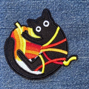 May include: A circular embroidered patch featuring a black cat with wide white eyes. The cat is curled around a piece of candy corn, which displays horizontal stripes of yellow, orange, and red. The patch has a black border and stitched details.