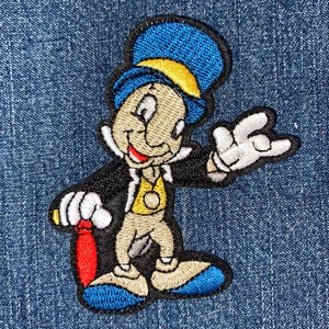 May include: Embroidered patch of a cartoon character wearing a blue top hat, a yellow vest, and holding a red stick. The character has a brown face, a big smile, and is wearing blue shoes.